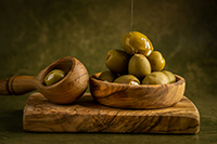 Olive oil being drizzled over a wooden bowl filled with olives.